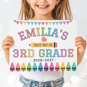 May include: A rectangular white sign featuring colourful bunting at the top and a row of crayons at the bottom. The text reads EMILIA'S FIRST DAY OF 3RD GRADE 2026-2027, with a red apple icon and a yellow pencil graphic centred on the sign.