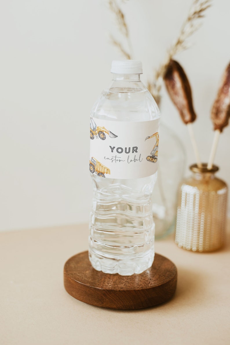 May include: A clear plastic water bottle with a white cap and a label that says "YOUR custom label". The label has a pattern of yellow construction vehicles. The bottle is sitting on a wooden coaster.