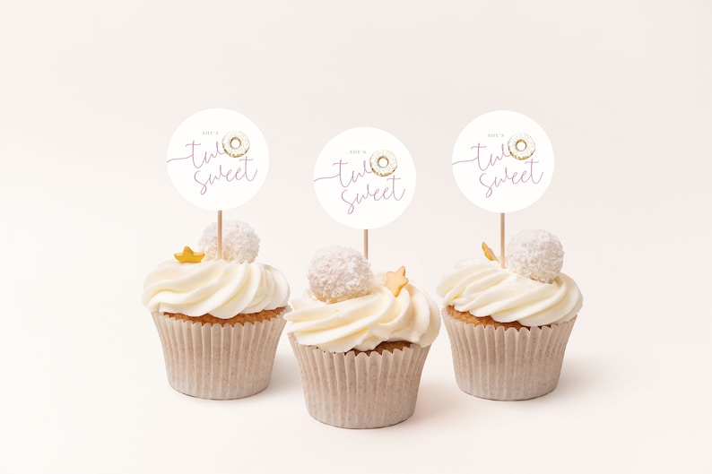 May include: Three white cupcakes with pink frosting and gold sprinkles. Each cupcake has a round white paper topper with a pink donut and the text "Donut Worry, Be Happy" in pink script.