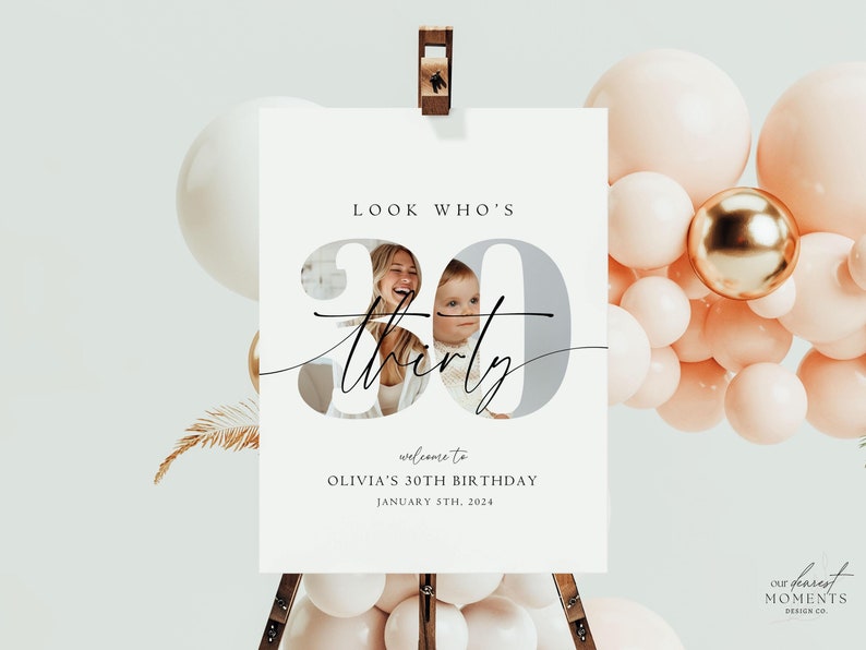 Op de afbeelding: Een wit bord met een foto van twee mensen in een grijs nummer 30 met de tekst "LOOK WHO'S 30 thirty" en "welcome to OLIVIA'S 30TH BIRTHDAY JANUARY 5TH, 2024". Het bord staat op een houten ezel met roze en gouden ballonnen erachter.