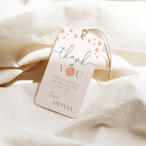 May include: Peach-themed thank you tag with the text "Thank You" and "For celebrating my special day with me. Love, Olivia". The tag is pink with peach illustrations and green leaves.