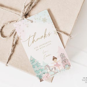 May include: A gift tag with a watercolor design featuring a ballerina, nutcracker, and rocking horse. The tag says "thanks" in gold script, with the text "for showering me with love" below. The tag is tied to a wrapped gift.