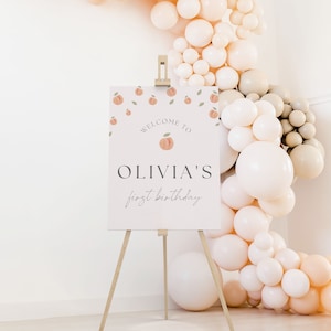 May include: A white welcome sign with peach illustrations and the text "Welcome to Olivia's first birthday". The sign is on a wooden easel in front of a wall decorated with peach and cream balloons.
