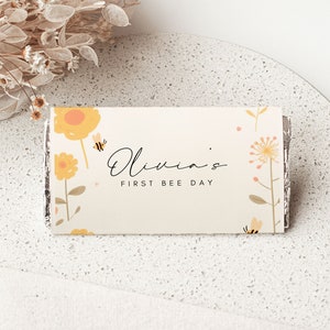 May include: A white card with a yellow and orange floral design and the text "Olivia's First Bee Day".