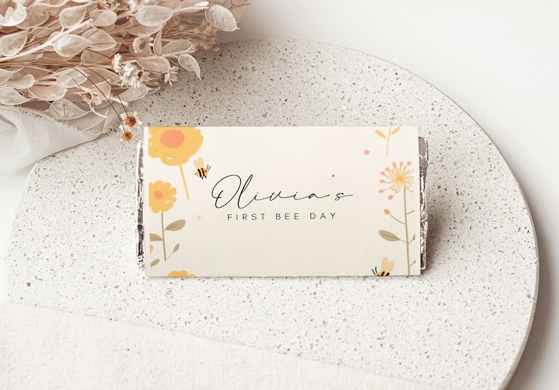 May include: A white card with a yellow and orange floral design and the text "Olivia's First Bee Day".