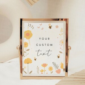 May include: A gold-framed picture with a white background and a floral design featuring yellow flowers, pink flowers, green leaves, and bees. The text "YOUR CUSTOM text" is written in black on the picture.