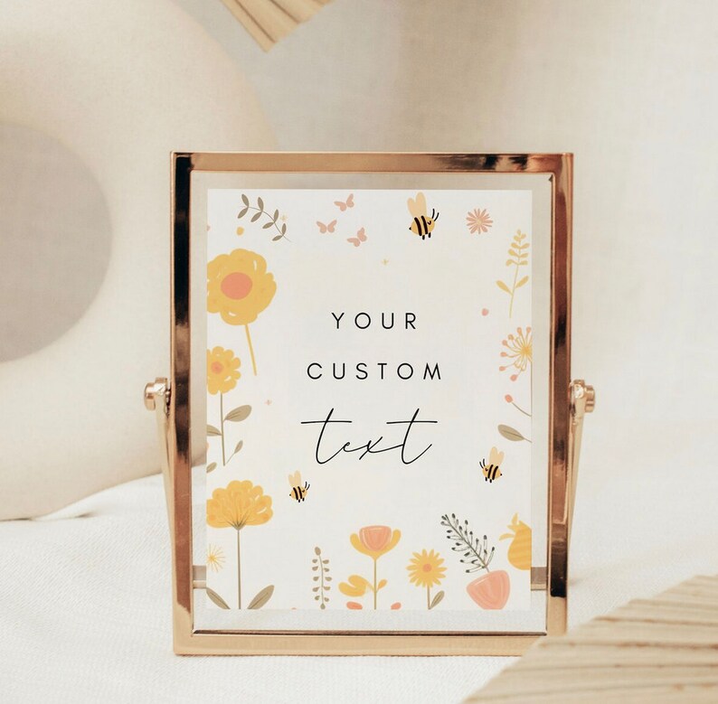 May include: A gold-framed picture with a white background and a floral design featuring yellow flowers, pink flowers, green leaves, and bees. The text "YOUR CUSTOM text" is written in black on the picture.