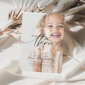 May include: A white invitation card with a photo of a young girl smiling. The card has a black and white design with the text "three" and "Chloe's Third Birthday".