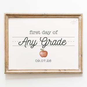Op de afbeelding: Een rechthoekige houten lijst met een wit bord waarop de zwarte tekst first day of Any Grade staat in een mix van typemachine- en cursieve lettertypes op gelinieerd papier. Een kleine rode aquarelappel staat onder de tekst met 09.07.26.