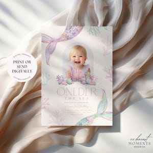 May include: A pink and blue mermaid-themed first birthday party invitation with a photo of a baby girl in a seashell crown. The invitation says "One-der the Sea" and includes the date, time, and location of the party.