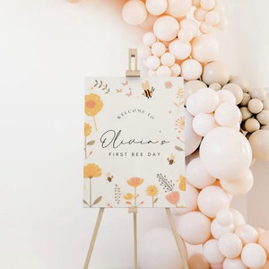May include: Welcome to Olivia's First Bee Day sign with a floral design featuring yellow flowers, bees, and butterflies. The sign is on a wooden easel and surrounded by a garland of cream and beige balloons.