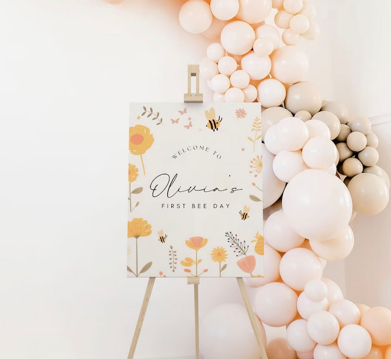 May include: Welcome to Olivia's First Bee Day sign with a floral design featuring yellow flowers, bees, and butterflies. The sign is on a wooden easel and surrounded by a garland of cream and beige balloons.