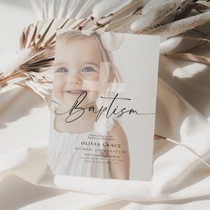 May include: A baptism invitation card featuring a smiling baby girl. The card has the word "Baptism" in elegant script, along with event details. The background includes dried flowers and soft fabric.