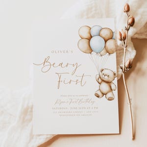 May include: A light brown teddy bear holding onto blue and brown balloons. The invitation reads "Oliver's Beary First" and includes the text "Please join us to celebrate Ryan's First Birthday, Saturday, June 26th at 4 PM, 121 Anywhere Street, Any City, Registered on Amazon". The bottom of the invitation reads "Our Dearest Moments Design Co."