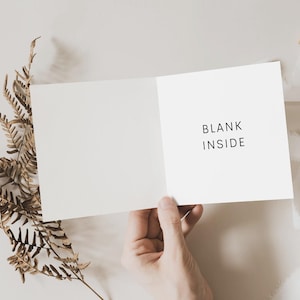 Pode incluir: Um cart&atilde;o branco em branco com o texto "BLANK INSIDE" impresso na frente. O cart&atilde;o est&aacute; sendo segurado aberto por uma m&atilde;o, e h&aacute; um ramo de samambaia marrom no fundo.