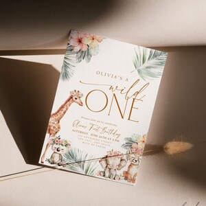 May include: A white invitation with a gold border and the text "Olivia's a wild ONE" in gold script. The invitation features a watercolor illustration of a giraffe, elephant, and koala with floral accents. The text "Please join us to celebrate Olivia's First Birthday" is printed in gold script. The invitation also includes the date, time, and location of the party.