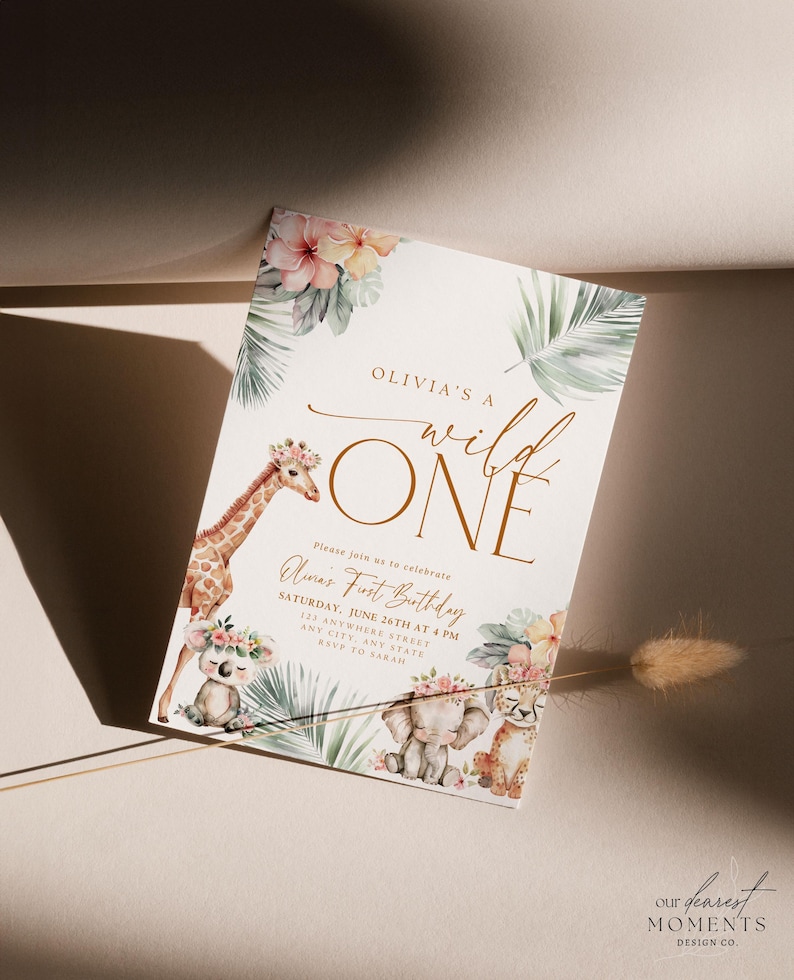 May include: A white invitation with a gold border and the text "Olivia's a wild ONE" in gold script. The invitation features a watercolor illustration of a giraffe, elephant, and koala with floral accents. The text "Please join us to celebrate Olivia's First Birthday" is printed in gold script. The invitation also includes the date, time, and location of the party.