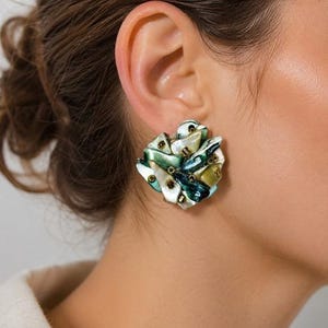May include: A cluster earring featuring iridescent, overlapping shell fragments in shades of deep teal, forest green, and creamy white. The organic, multi-layered design is accented with small, dark metallic beads interspersed throughout the surface.