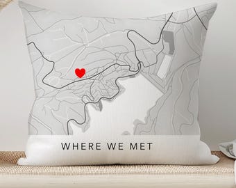 Puede incluir: Un cojín decorativo con un diseño de mapa en tonos grises, con un corazón rojo que marca una ubicación. El texto "WHERE WE MET" está impreso en negro en la parte inferior del cojín. El cojín está colocado sobre un banco de madera clara.