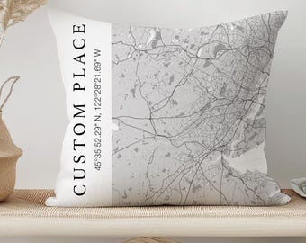 Puede incluir: Un cojín decorativo con un diseño de mapa personalizado en tonos grises. El cojín presenta el texto "CUSTOM PLACE" y las coordenadas 45°35'52.29" N, 122°28'21.69" W. El cojín está colocado en un banco de madera clara.
