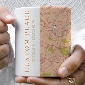Op de afbeelding: Witte keramische mok met een aangepast kaartontwerp. De mok heeft de tekst "CUSTOM PLACE" in een verticale, oranje lettertype, samen met coördinaten. Het kaartontwerp is in tinten van bruin, bruin en groen.