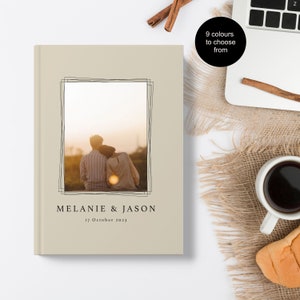 May include: A beige photo album with a black frame around a photo of a couple standing in a field at sunset. The album has the text "Melanie & Jason" and "17 October 2023" printed on the front.