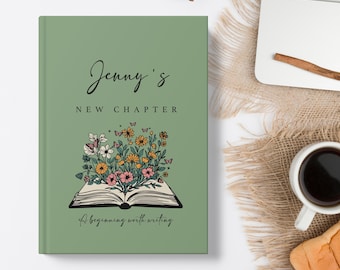 Puede incluir: Un diario verde salvia con el texto "Jenny's New Chapter" y una ilustración de un libro abierto con flores y mariposas. La frase "A beginning worth writing" está en la parte inferior. El diario está sobre una superficie blanca.