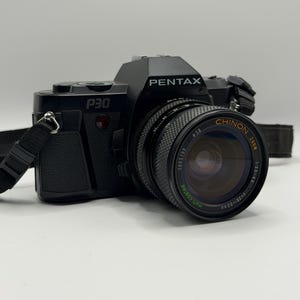 Peut inclure: Un appareil photo reflex argentique 35mm Pentax P30 noir équipé d'un objectif zoom Chinon avec une ouverture de 1:3.5-4.5 et une focale de 28-50mm. Le boîtier comprend une sangle de cou noire fixée aux attaches latérales et le logo Pentax.