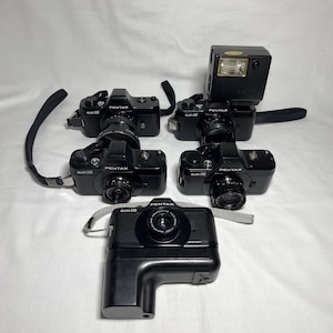 May include: A collection of five black Pentax cameras, including a camera with a flash attachment. The cameras are arranged on a white surface. Each camera has a lens and various buttons and dials. Some cameras have straps.