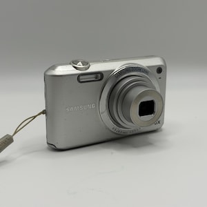 Pode incluir: Uma câmara digital Samsung prateada com lente de zoom retrátil, flash integrado e corpo metálico. A parte frontal do dispositivo exibe a marca Samsung e as especificações da lente, incluindo uma correia de pulso em cinzento claro.