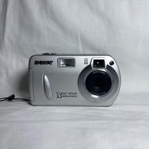 Könnte beinhalten: Eine silberne Sony Cyber-shot Digitalkamera mit einer Auflösung von 3,2 Megapixel und einem 7,5 mm Objektiv. Die Kamera hat einen schwarzen Gurt.