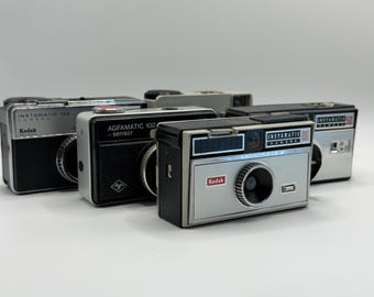 May include: A collection of four vintage film cameras including a Kodak Instamatic 133, an Agfamatic 100 sensor, a Kodak Instamatic 100, and a Kodak Instamatic 50. These rectangular cameras feature black and silver metal bodies with various brand labels.
