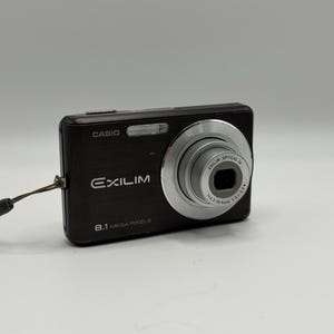 Puede incluir: Una cámara digital Casio Exilim negra con un sensor de 8,1 megapíxeles, un objetivo plateado y un flash integrado. La parte frontal muestra la marca Casio, el modelo Exilim y una correa de muñeca marrón sujeta a un lateral.