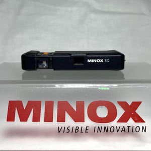 以下が含まれることがあります： 黒いMinox ECカメラ、コンパクトフィルムカメラが透明な表面に展示されています。カメラは長方形で、前面に「MINOX EC」の文字が印刷されています。「MINOX」のブランド名は赤色で表示されています。