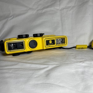 Pode incluir: Câmera subaquática Minolta amarela com alça de pulso preta. A câmera tem formato retangular com lente, flash e mostradores pretos. O logotipo da Minolta é visível.