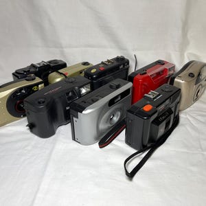 May include: A collection of eight vintage 35mm film cameras in various colors including black, silver, gold, and red. The cameras feature rectangular bodies, built-in flashes, and viewfinders, with some models displaying brand names like Kodak.