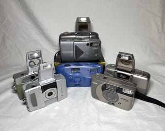 May include: A collection of vintage point-and-shoot film cameras in various colors, including silver, blue, and gray. The cameras are arranged on a white surface, showcasing their different designs and features. Some have pop-up flashes.