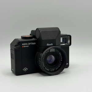 May include: A black Agfa Optima Sensor camera featuring a prominent lens with text reading Agfa Solitar 1:2.8/40. The camera body includes a flash unit on top, a shutter button with an orange dot, and the Agfa logo on the front face of the device.