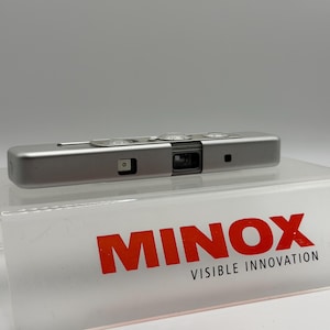 May include: A silver rectangular Minox subminiature camera featuring a central lens, a viewfinder, and a flash sync port. The camera is positioned on a clear acrylic display stand with the red text MINOX VISIBLE INNOVATION printed on the front.