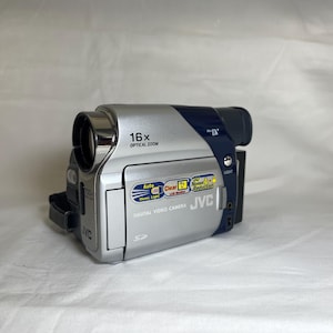Puede incluir: Videocámara digital JVC plateada y azul marino con zoom óptico de 16x. Incluye luz automática, monitor LCD claro y reducción de ruido. Incluye una ranura para tarjeta SD y el logotipo de JVC.