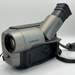 May include: A gray Samsung camcorder featuring a lens with 88x digital zoom, a high-resolution viewfinder, and a black carrying strap. The device has a compact, handheld design with a prominent lens barrel and a side-mounted battery compartment.