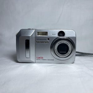 May include: Silver Olympus C-500 Zoom digital camera. The camera has a built-in flash, a lens with the text "Olympus Lens", and a wrist strap. The camera has the text "5.0 Megapixels" and "Camedia" printed on it.