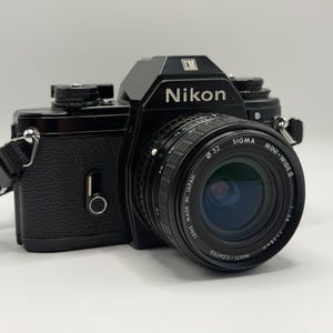 May include: A black Nikon EM 35mm film camera featuring a textured body and a black Sigma Mini-Wide II 28mm f/2.8 lens. The lens barrel displays the text Sigma Mini-Wide II, 52mm filter diameter, and Lens Made in Japan. A black strap is attached.