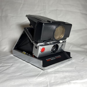 Op de afbeelding: Een zilveren Polaroid SX-70 Land Camera met een zwarte lens en een rode knop. De camera is open en de flitser is zichtbaar.