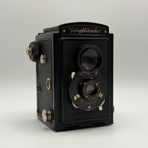 Könnte beinhalten: Eine schwarze Voigtländer Brillant Kamera mit Doppellinsen-Design. Die Kamera hat einen rechteckigen Körper mit silberfarbenen Knöpfen und einem Objektiveinstellrad. Das Wort "Voigtländer" ist in goldener Schrift aufgedruckt.