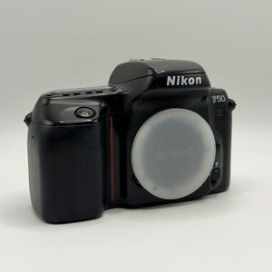 May include: A black Nikon F50 35mm film SLR camera body featuring a white circular lens cap with the Nikon logo. The camera has a prominent viewfinder hump, a shutter button on the grip, and a red vertical accent line on the front panel.