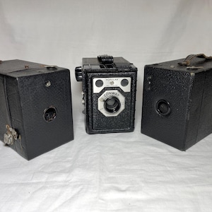 May include: Three vintage box cameras with black textured bodies. The centre camera features a silver faceplate with the text "POPULAR" and "CONWAY" printed above the lens. Each camera has a rectangular shape, a central lens, and a top handle.