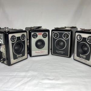 Könnte beinhalten: Vier Vintage-Boxkameras von Kodak Brownie, die in einer Reihe angeordnet sind. Die Kameras sind schwarz mit silbernen Frontplatten und verfügen über kreisförmige Linsen und Sucher. Der Text umfasst Brownie Flash II, Model E, Model C und Model D.