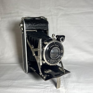 May include: A vintage black and silver folding camera with a lens and viewfinder. The camera has a bellows design and a silver metal frame. The lens has text around the edge. The camera is open and ready for use.