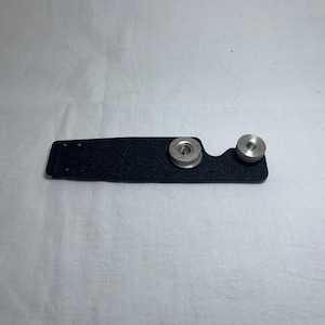 Puede incluir: Un accesorio de cámara negro y rectangular con dos perillas circulares plateadas. El accesorio tiene una superficie texturizada y está sobre un fondo blanco. Las perillas son de diferentes tamaños.